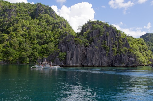 A traditional boat floats on clear, calm waters near a large limestone cliff covered in lush green vegetation. The sky is bright with scattered white clouds, adding to the scenic beauty of the tropical landscape.