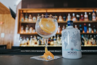 A glass garnished with an orange slice and peel holds a clear beverage on a bar counter, with a bottle of Nordés Atlantic Galician Gin placed beside it. Behind the counter, shelves display various bottles, softly illuminated in warm light.
