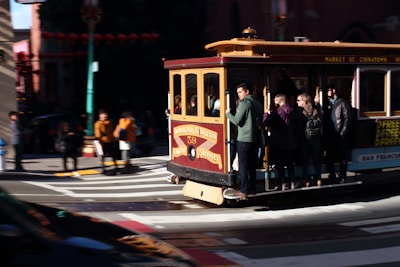 A classic cable car carrying several passengers travels down a street in an urban area, with people visible in the background. The cable car has a traditional design with vivid colors, and some passengers are standing and holding onto the rails.
