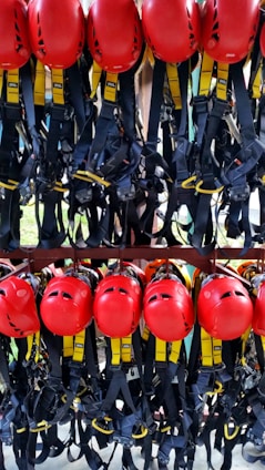 A collection of bright red helmets and black harnesses with yellow accents are neatly organized and hanging on a rack. They are likely used for safety in activities such as climbing or construction.