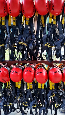 A collection of bright red helmets and black harnesses with yellow accents are neatly organized and hanging on a rack. They are likely used for safety in activities such as climbing or construction.