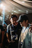 Two individuals wearing name badges and lanyards are engaged in a close conversation indoors. One person has their arm around the other's shoulder, suggesting camaraderie or support. The lighting is warm and diffused, possibly from an overhead source, and the background features a draped ceiling with circular light patterns.