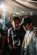 Two individuals wearing name badges and lanyards are engaged in a close conversation indoors. One person has their arm around the other's shoulder, suggesting camaraderie or support. The lighting is warm and diffused, possibly from an overhead source, and the background features a draped ceiling with circular light patterns.