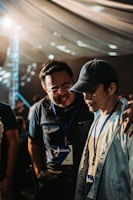 Two individuals wearing name badges and lanyards are engaged in a close conversation indoors. One person has their arm around the other's shoulder, suggesting camaraderie or support. The lighting is warm and diffused, possibly from an overhead source, and the background features a draped ceiling with circular light patterns.