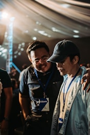 Two individuals wearing name badges and lanyards are engaged in a close conversation indoors. One person has their arm around the other's shoulder, suggesting camaraderie or support. The lighting is warm and diffused, possibly from an overhead source, and the background features a draped ceiling with circular light patterns.