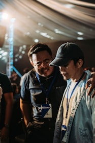 Two individuals wearing name badges and lanyards are engaged in a close conversation indoors. One person has their arm around the other's shoulder, suggesting camaraderie or support. The lighting is warm and diffused, possibly from an overhead source, and the background features a draped ceiling with circular light patterns.