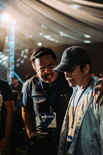 Two individuals wearing name badges and lanyards are engaged in a close conversation indoors. One person has their arm around the other's shoulder, suggesting camaraderie or support. The lighting is warm and diffused, possibly from an overhead source, and the background features a draped ceiling with circular light patterns.