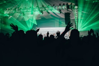 Silhouettes of people with raised hands are in the foreground, set against a backdrop of vibrant green stage lights and a metal truss supporting various lights. A partially visible screen displays text amidst the celebratory atmosphere, under a large tent-like ceiling.