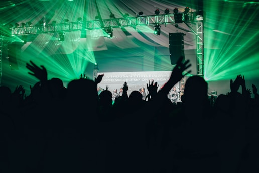 Silhouettes of people with raised hands are in the foreground, set against a backdrop of vibrant green stage lights and a metal truss supporting various lights. A partially visible screen displays text amidst the celebratory atmosphere, under a large tent-like ceiling.
