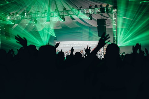Silhouettes of people with raised hands are in the foreground, set against a backdrop of vibrant green stage lights and a metal truss supporting various lights. A partially visible screen displays text amidst the celebratory atmosphere, under a large tent-like ceiling.