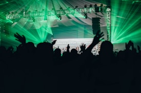 Silhouettes of people with raised hands are in the foreground, set against a backdrop of vibrant green stage lights and a metal truss supporting various lights. A partially visible screen displays text amidst the celebratory atmosphere, under a large tent-like ceiling.