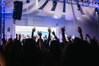 A large group of people with their hands raised in the air inside a dimly lit auditorium. There are stage lights and a backdrop with text partly visible in the background. Drapes hang from the ceiling, adding to the event atmosphere.