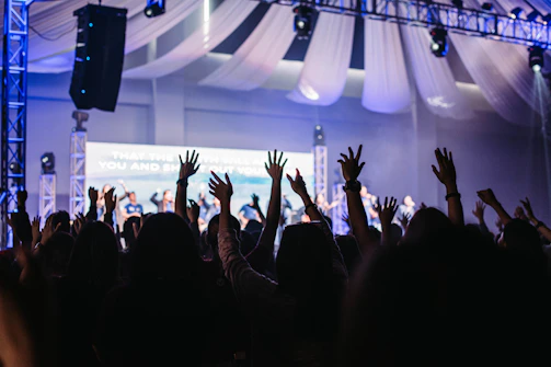A large group of people with their hands raised in the air inside a dimly lit auditorium. There are stage lights and a backdrop with text partly visible in the background. Drapes hang from the ceiling, adding to the event atmosphere.