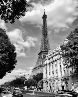 A black and white photograph of a cityscape featuring a prominent tower with a lattice design against a partly cloudy sky. Tree foliage frames the image, and a street with cars and pedestrians is visible. Classic architecture lines the street, showcasing a blend of nature and urban elements.