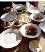 Various breakfast items are arranged on a wooden table, including sliced bread, cheese, pastries, sausages, and a meat dish, accompanied by empty plates and glasses. The sunlight is casting shadows on the table, enhancing the warm atmosphere of the setting.