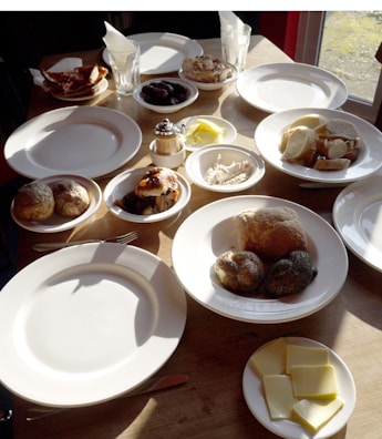 Various breakfast items are arranged on a wooden table, including sliced bread, cheese, pastries, sausages, and a meat dish, accompanied by empty plates and glasses. The sunlight is casting shadows on the table, enhancing the warm atmosphere of the setting.