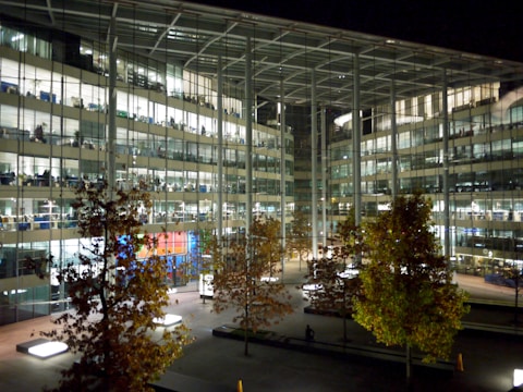 A modern glass office building at night, with multiple floors brightly lit from within. The interior is visible through the large glass windows, showcasing desks, chairs, and computers. In front of the building, several trees with autumn-colored leaves are present, and the courtyard includes lit benches and pathways.