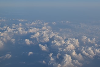 A vast expanse of soft, fluffy clouds viewed from above, merging with a clear, light blue sky. The light subtly highlights the cloud contours, creating a serene and tranquil atmosphere.