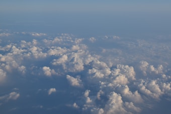 A vast expanse of soft, fluffy clouds viewed from above, merging with a clear, light blue sky. The light subtly highlights the cloud contours, creating a serene and tranquil atmosphere.