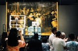 A large, historical painting is displayed in a museum setting with a crowd gathered in front of it. The painting is housed in a protective structure with visible machinery and equipment. People are taking photos and engaging with the exhibition.