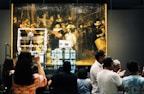 A large, historical painting is displayed in a museum setting with a crowd gathered in front of it. The painting is housed in a protective structure with visible machinery and equipment. People are taking photos and engaging with the exhibition.