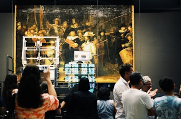 A large, historical painting is displayed in a museum setting with a crowd gathered in front of it. The painting is housed in a protective structure with visible machinery and equipment. People are taking photos and engaging with the exhibition.