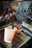 A coffee machine is dispensing espresso into a paper cup. The machine features a metal portafilter and a series of buttons for different functions. The scene captures the fresh coffee flowing from the spout into the cup positioned below.