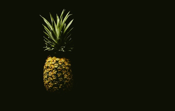A pineapple is set against a dark background, creating a dramatic and striking contrast. The lighting highlights the texture of the fruit's rough outer skin and the spiky green leaves atop it.