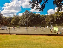 A group of people are playing tennis on an outdoor court. The players appear to be engaged in various activities, possibly part of a tennis lesson or practice session. The tennis court is surrounded by a fence and there are trees in the background. The sky is blue with some fluffy white clouds. Equipment and chairs can be seen along the sides of the court.