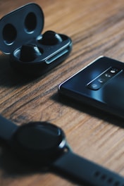 A set of black wireless earbuds in an open charging case is placed on a wooden surface. Nearby, there is a sleek smartphone with a multi-lens camera array facing upwards. In the foreground, a smart watch with a black band lies nearby, slightly out of focus.
