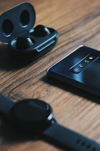 A set of black wireless earbuds in an open charging case is placed on a wooden surface. Nearby, there is a sleek smartphone with a multi-lens camera array facing upwards. In the foreground, a smart watch with a black band lies nearby, slightly out of focus.