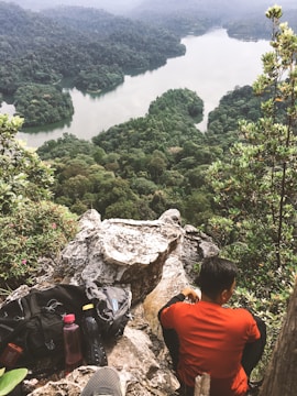 A picturesque landscape featuring a dense forest surrounding a tranquil lake. A person in a red shirt is sitting on a rocky outcrop, looking out over the scene. Nearby, a backpack and water bottle are placed on the rocks, suggesting a hiking or trekking excursion. The tall trees frame the view, adding a sense of serene isolation.