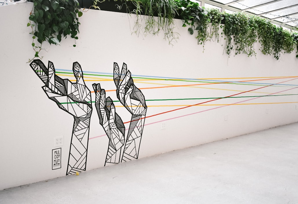 Mural of three geometric hands with rainbow strings coming from them, across a wall topped with ivy