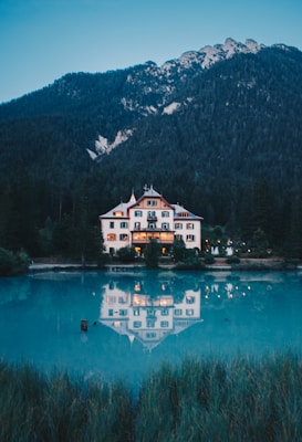 A large, elegant house with multiple windows and a steep roof is nestled at the base of a forested mountain. The house is beautifully reflected in the calm, turquoise lake in front of it. The entire scene is enveloped in an evening or early morning light, providing a serene and tranquil atmosphere.