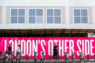 A group of people walk past a vivid pink sign with bold white text that reads 'LONDON'S OTHER SIDE'. In front of the sign, a row of bicycles is parked in a docking station. The background includes a building with a white facade and several windows.