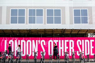 A group of people walk past a vivid pink sign with bold white text that reads 'LONDON'S OTHER SIDE'. In front of the sign, a row of bicycles is parked in a docking station. The background includes a building with a white facade and several windows.