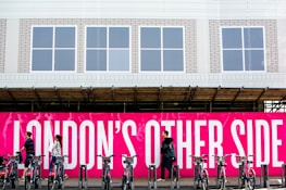 A group of people walk past a vivid pink sign with bold white text that reads 'LONDON'S OTHER SIDE'. In front of the sign, a row of bicycles is parked in a docking station. The background includes a building with a white facade and several windows.