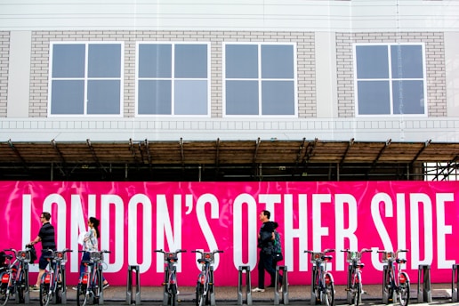 A group of people walk past a vivid pink sign with bold white text that reads 'LONDON'S OTHER SIDE'. In front of the sign, a row of bicycles is parked in a docking station. The background includes a building with a white facade and several windows.