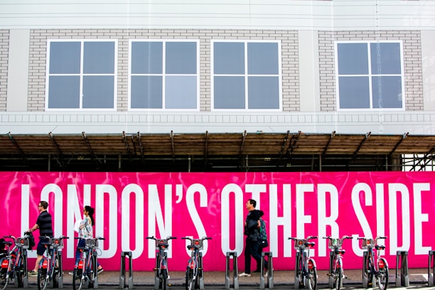 A group of people walk past a vivid pink sign with bold white text that reads 'LONDON'S OTHER SIDE'. In front of the sign, a row of bicycles is parked in a docking station. The background includes a building with a white facade and several windows.