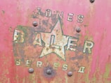 A close-up of an old, rusty metal surface displaying faded lettering and a large star logo. The words 'JONES BALER SERIES II' are partially visible amidst patches of oxidation and weathering. The surface shows a mix of red, green, and brown hues, hinting at long-term exposure to the elements.