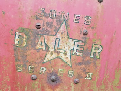 A close-up of an old, rusty metal surface displaying faded lettering and a large star logo. The words 'JONES BALER SERIES II' are partially visible amidst patches of oxidation and weathering. The surface shows a mix of red, green, and brown hues, hinting at long-term exposure to the elements.