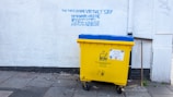 A large yellow and blue waste bin is placed on a concrete sidewalk near a building with a white textured wall. Above the bin, there is graffiti text in blue, offering various used items such as bicycles and DVDs, and mentioning a shop named 'The Three Divine Virtues' Shop.' The wall has a pipe running vertically along its surface. The bin has a label indicating that no food waste is allowed.
