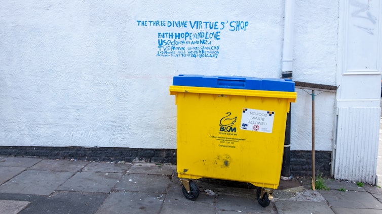 A large yellow and blue waste bin is placed on a concrete sidewalk near a building with a white textured wall. Above the bin, there is graffiti text in blue, offering various used items such as bicycles and DVDs, and mentioning a shop named 'The Three Divine Virtues' Shop.' The wall has a pipe running vertically along its surface. The bin has a label indicating that no food waste is allowed.