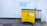 A large yellow and blue waste bin is placed on a concrete sidewalk near a building with a white textured wall. Above the bin, there is graffiti text in blue, offering various used items such as bicycles and DVDs, and mentioning a shop named 'The Three Divine Virtues' Shop.' The wall has a pipe running vertically along its surface. The bin has a label indicating that no food waste is allowed.