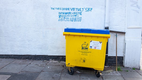 A large yellow and blue waste bin is placed on a concrete sidewalk near a building with a white textured wall. Above the bin, there is graffiti text in blue, offering various used items such as bicycles and DVDs, and mentioning a shop named 'The Three Divine Virtues' Shop.' The wall has a pipe running vertically along its surface. The bin has a label indicating that no food waste is allowed.