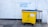 A large yellow and blue waste bin is placed on a concrete sidewalk near a building with a white textured wall. Above the bin, there is graffiti text in blue, offering various used items such as bicycles and DVDs, and mentioning a shop named 'The Three Divine Virtues' Shop.' The wall has a pipe running vertically along its surface. The bin has a label indicating that no food waste is allowed.