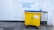 A large yellow and blue waste bin is placed on a concrete sidewalk near a building with a white textured wall. Above the bin, there is graffiti text in blue, offering various used items such as bicycles and DVDs, and mentioning a shop named 'The Three Divine Virtues' Shop.' The wall has a pipe running vertically along its surface. The bin has a label indicating that no food waste is allowed.