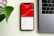 A smartphone displaying subscriber statistics with a rising graph and specific numbers on a red background sits on a desk beside a silver laptop. A green plant leaf is partially visible on the left side of the image.