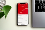 A smartphone displaying subscriber statistics with a rising graph and specific numbers on a red background sits on a desk beside a silver laptop. A green plant leaf is partially visible on the left side of the image.