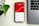 A smartphone displaying subscriber statistics with a rising graph and specific numbers on a red background sits on a desk beside a silver laptop. A green plant leaf is partially visible on the left side of the image.